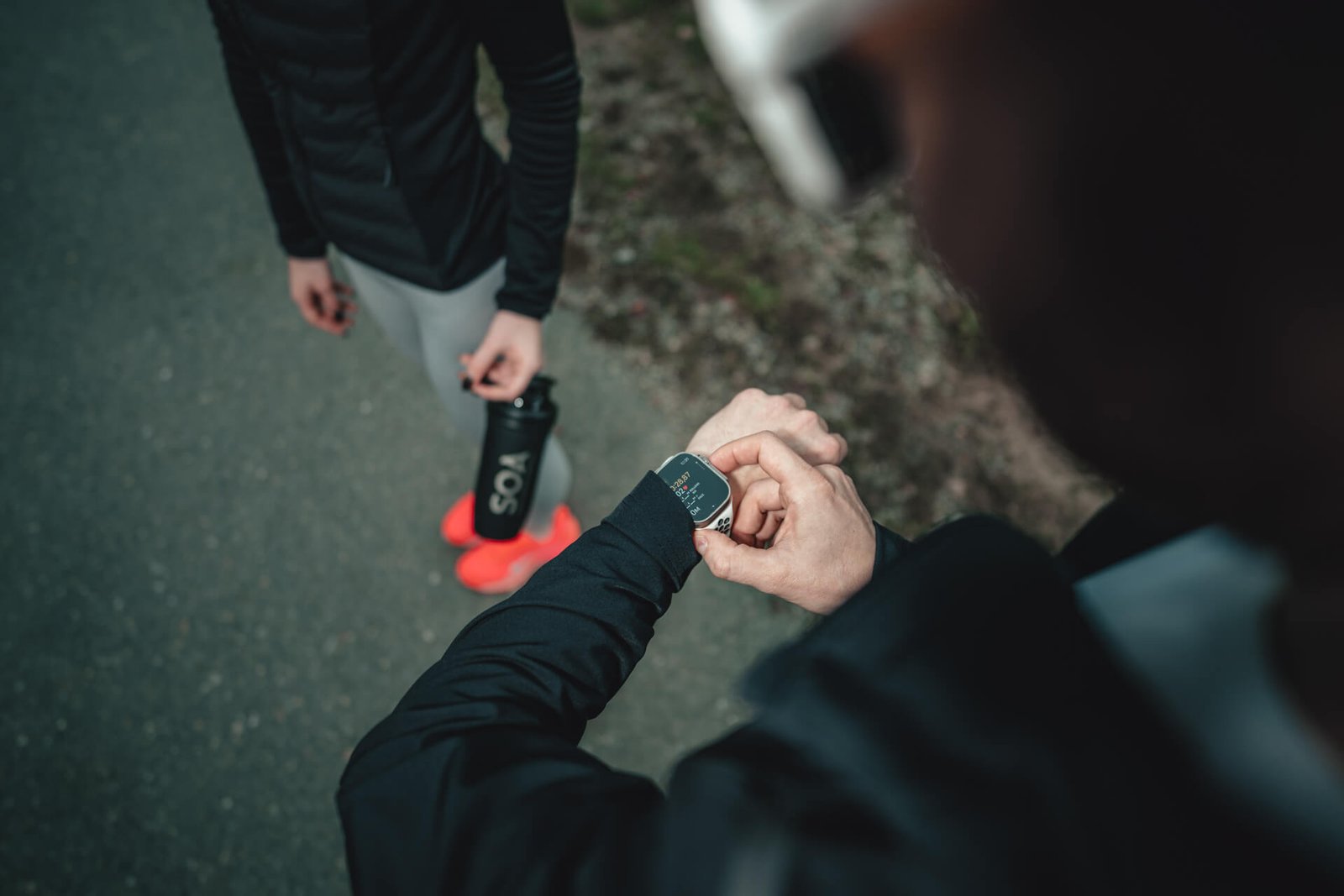 SOA Running – Sportler checkt Trainingszeit auf Laufuhr, Partnerin mit Shaker im Hintergrund